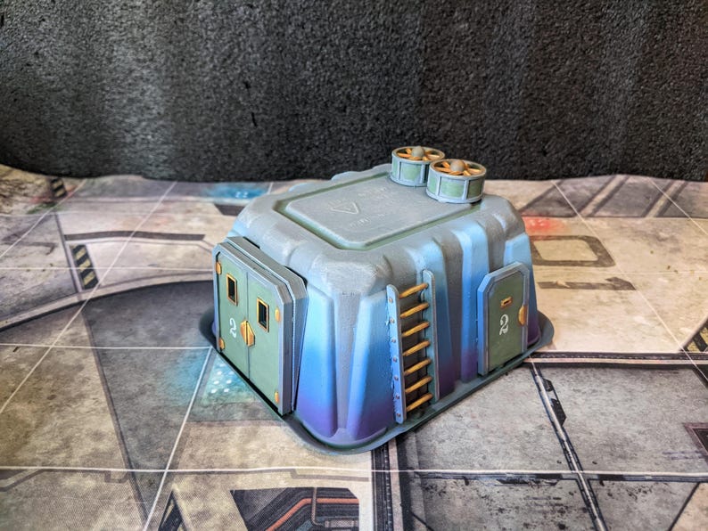 May include: A miniature, futuristic-style building with a gray and blue color scheme. The structure features two small ventilation units on top, two green doors with the number "2", and a small orange ladder. The building is set on a patterned surface.