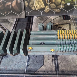 May include: A set of gray and yellow plastic barriers for a tabletop game. The barriers are in various shapes and sizes, including straight, curved, and angled pieces. Some of the barriers have a skull and crossbones design, while others have a smiley face design.