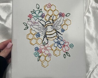 May include: Embroidered canvas art featuring a bee surrounded by flowers and a honeycomb pattern. The bee is yellow and black, the flowers are pink, blue, and green, and the honeycomb is gold.