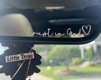 May include: A car rearview mirror with the words "Trust in God" in a cursive font, with a heart symbol. A black air freshener with the words "Little Trees" and "Black Ice" hangs below the mirror.