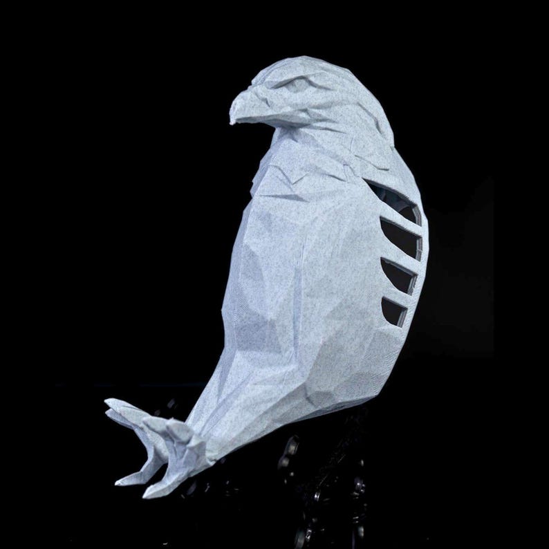 May include: A light gray, geometric paper sculpture of a bird of prey, possibly an eagle or hawk, with a detailed head and open wing design. The artwork is set against a black background, highlighting the paper's texture and form.