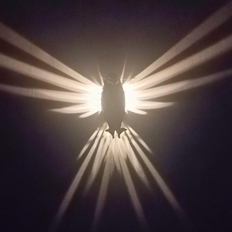 May include: A wall-mounted owl-shaped light fixture. The fixture casts a pattern of light and shadow resembling wings. The owl is silhouetted against the light, with the light radiating outwards in a fan-like pattern. The background is a dark color.