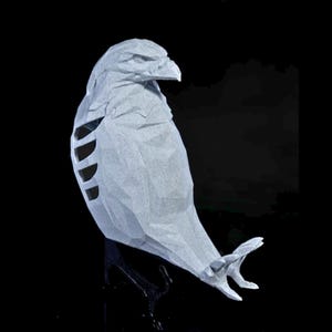 May include: A light gray paper sculpture of a bird, possibly an owl or hawk, with detailed feather and wing designs. The bird is perched on a black stand against a black background. The sculpture has a textured appearance.
