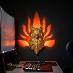 Könnte beinhalten: Eine goldfarbene, geometrische Fuchskopf-Wandleuchte mit orangefarbenem Schein, der von hinten ausgeht. Die Leuchte ist an einer dunklen Wand über einem Schreibtisch mit Tastatur und Computermonitor angebracht.
