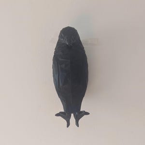 May include: A black, geometric-shaped bird sculpture mounted on a white wall. The bird has a textured surface and is attached with clear tape. The sculpture's design features sharp angles and a stylized form.