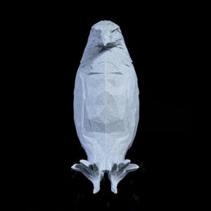 May include: A light gray, geometric origami sculpture of a bird, possibly an owl or eagle, against a black background. The sculpture has a polygonal design, with visible facets creating a textured appearance. The bird's form is upright, with its wings folded.
