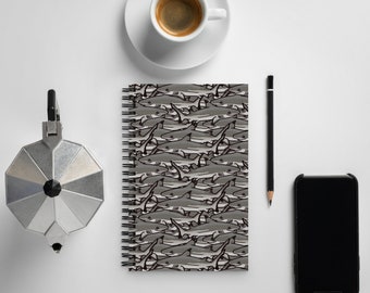 May include: A spiral bound notebook with a gray and white shark pattern. The notebook is on a white surface with a coffee cup, a pencil, and a smartphone.