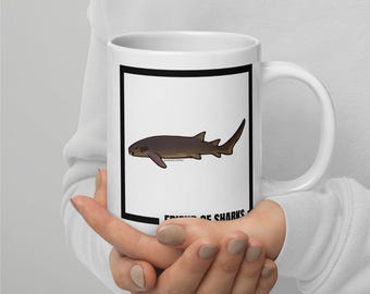 May include: White ceramic mug featuring a brown shark illustration within a black border. The mug has a curved handle and the text "FRIENDS OF SHARKS" at the bottom. The shark design is detailed with fins and a tail.
