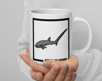 May include: White ceramic mug featuring a gray cartoon shark with a long tail, set against a white background within a black border. The text "FRIEND OF SHARKS" is printed below the shark illustration.