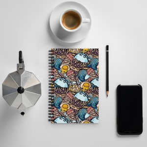 Puede incluir: Una libreta de espiral con un diseño colorido de estilo de dibujos animados que presenta medusas, huevos y otras formas abstractas. La libreta está sobre una superficie blanca con una taza de café, un lápiz y un teléfono inteligente.