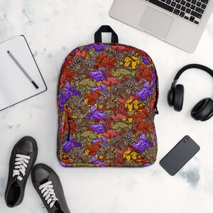 Pode incluir: Uma mochila com um padrão colorido de peixes em tons de vermelho, roxo, amarelo e castanho. A mochila tem um fecho de correr e uma pega pretos. Está sobre uma superfície branca com um laptop, auscultadores e um smartphone.