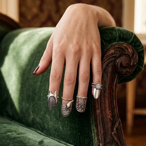 May include: A set of four silver-toned nail rings worn on the fingers. Each ring features a unique design, including one with a textured surface, one with a small animal motif, one with a floral pattern, and one with a smooth, polished finish.