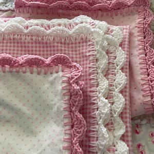 May include: A stack of pink and white crocheted fabric squares with different patterns. The fabric squares are layered on top of each other, with the pink squares on top. The fabric squares are all edged with crocheted trim.