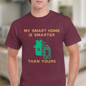 Puede incluir: Una camiseta color granate con el texto "My smart home is smarter than yours" en amarillo y un gráfico verde de una casa con un teléfono inteligente y un diseño de placa de circuito.