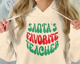 Könnte beinhalten: Ein cremefarbener Hoodie mit einer grünen und roten Grafik, auf der in einer Retro-Schrift "Santas Lieblingslehrer" steht.