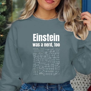 Puede incluir: Una sudadera azul turquesa con el texto "Einstein era un nerd, también" en letras blancas. El texto está rodeado por un gráfico blanco de ecuaciones matemáticas.