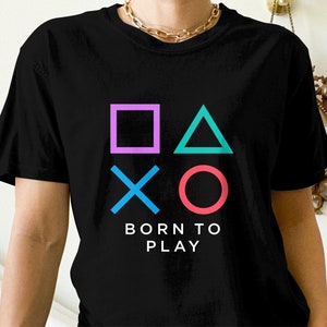 May include: Black t-shirt featuring a graphic of the PlayStation controller symbols in pink, blue, green, and red. The text "Born to Play" is printed below the symbols.