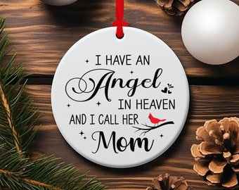 Op de afbeelding: Een witte keramische kerstversiering met de tekst "I HAVE AN Angel IN HEAVEN AND I CALL HER Mom". Een rode kardinaal zit op een tak. De versiering hangt aan een rood lint, omringd door dennennaalden, dennenappels en zilveren ballen.
