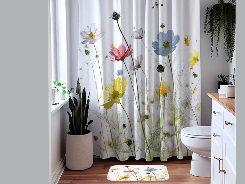 May include: A white shower curtain with a floral print featuring colourful cosmos flowers on a white background. The shower curtain is in a bathroom with a white toilet and a white cabinet with gold hardware.