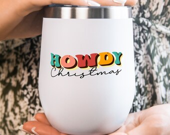 以下が含まれることがあります： 白いステンレス製のタンブラーに、カラフルな「Howdy Christmas」のグラフィックが描かれています。タンブラーは手で持たれています。