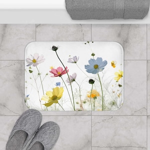 May include: A white bath mat with a floral design featuring pink, yellow, blue, and white flowers. The flowers are on white stems against a white background.