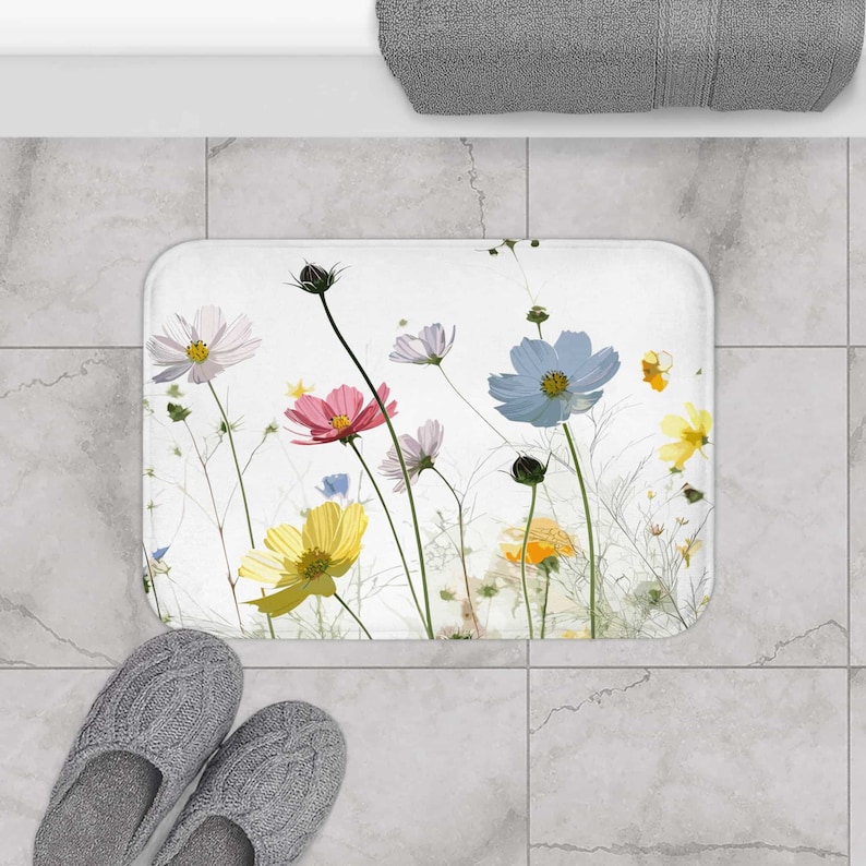 May include: A white bath mat with a floral design featuring pink, yellow, blue, and white flowers. The flowers are on white stems against a white background.