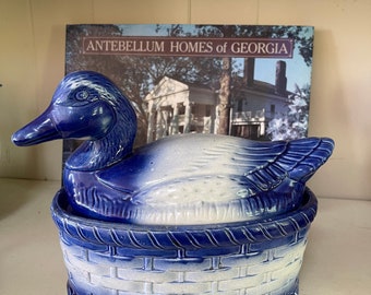 May include: A blue and white ceramic duck-shaped covered dish. The duck is sitting on a basket-shaped base. The dish is decorated with a blue and white pattern. The text "ANTEBELLUM HOMES of GEORGIA" is visible on a book in the background.