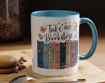 May include: White ceramic mug with a blue interior and handle. The mug features the text "Take me to the Bookstore" with floral and celestial accents. A row of illustrated books decorates the lower portion of the mug.