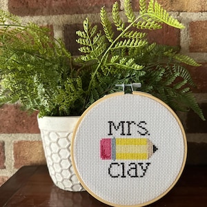 May include: A cross-stitch embroidery hoop with the text "Mrs. Clay" and a yellow pencil design with pink and black accents. The embroidery is set against a white background and framed in a wooden hoop. A potted plant is in the background.