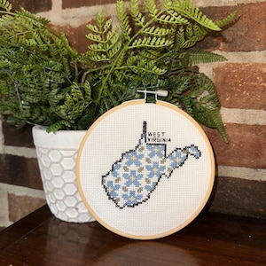 May include: A cross-stitch embroidery hoop featuring the state of West Virginia in blue, gray, and white. The text "WEST VIRGINIA" is stitched above the state outline. The hoop is set against a brick wall and a white pot with green fern.