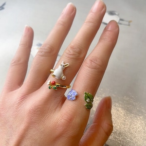 Puede incluir: Dos anillos de oro con detalles de esmalte. Un anillo presenta un conejo blanco con una perla y hojas verdes. El otro anillo presenta una flor azul con hojas verdes.