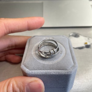 Op de afbeelding: Een zilveren ring met een delicaat ontwerp met meerdere banden en kleine glinsterende stenen. De ring wordt gepresenteerd in een grijze fluwelen ringdoos.