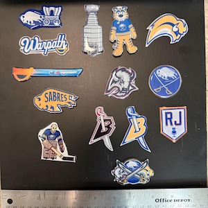May include: A collection of colorful enamel pins featuring various logos and designs. Includes a Stanley Cup, a tiger mascot, and logos with the words "Warpath" and "Sabres". The pins are displayed on a black surface with a ruler.