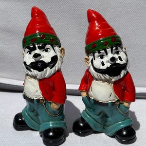 May include: Two ceramic garden gnomes with red hats, green and red striped headbands, and red jackets. The gnomes have white beards and mustaches, blue jeans, and black shoes. They are standing with their hands in their pockets.