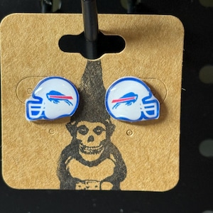 May include: A pair of stud earrings featuring a white and blue football helmet design with a red stripe and a buffalo logo. The earrings are displayed on a brown card with a black background.