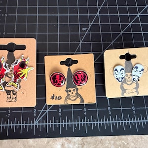 May include: Three sets of stud earrings displayed on brown cardboard backing cards. The left set features red and yellow hooded figures. The center set shows red circular icons with black silhouettes. The right set depicts white clown faces with black markings.
