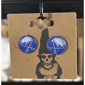 May include: A pair of round stud earrings with a blue background and a white graphic of a ram and crossed swords. The earrings are displayed on a brown cardboard backing with a black and white gnome illustration.
