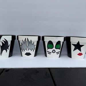 May include: A set of four cylindrical plant pots painted with black and white designs inspired by rock band makeup. Each pot features a unique graphic pattern, including black star shapes, green eye details, and red lips on a white and black background.