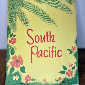 Puede incluir: Un folleto amarillo con hojas de palmera verdes y flores rojas alrededor del borde. El título "South Pacific" está escrito en letras rojas.