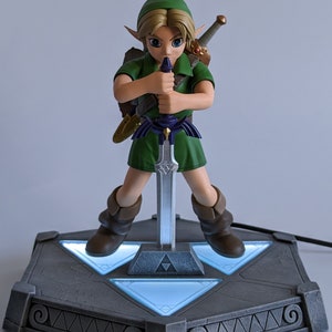 May include: A green-clad Link figurine from The Legend of Zelda series, holding the Master Sword, stands on a gray, triangular base with glowing blue lights. The base features a Triforce symbol and intricate details.