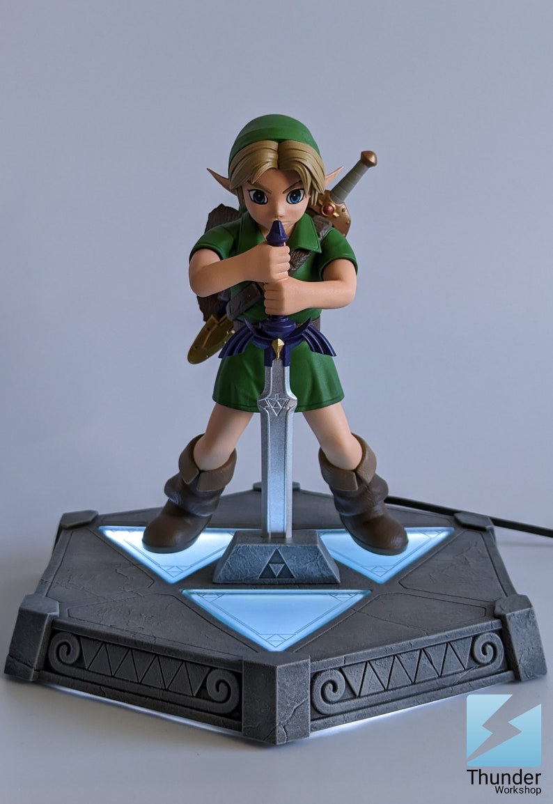 May include: A green-clad Link figurine from The Legend of Zelda series, holding the Master Sword, stands on a gray, triangular base with glowing blue lights. The base features a Triforce symbol and intricate details.