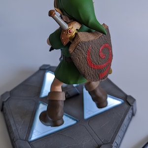 May include: A green and brown figurine of Link from The Legend of Zelda video game series. The figurine is standing on a gray base with a glowing blue Triforce symbol. The figurine is holding a sword and shield.