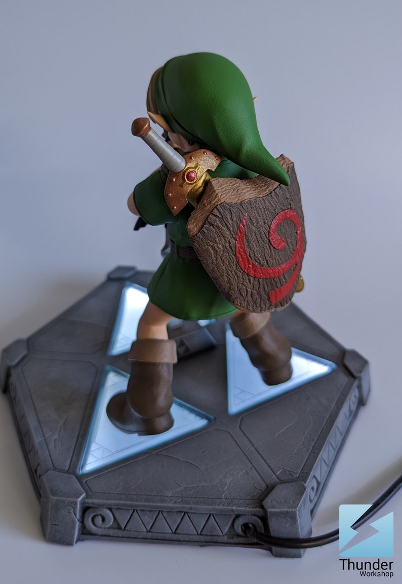 May include: A green and brown figurine of Link from The Legend of Zelda video game series. The figurine is standing on a gray base with a glowing blue Triforce symbol. The figurine is holding a sword and shield.