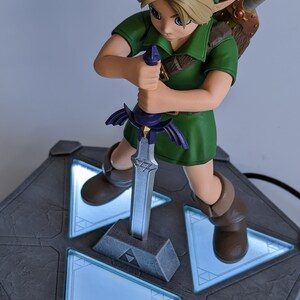 May include: A green-clad Link figurine from The Legend of Zelda: Ocarina of Time, holding the Master Sword, stands on a gray base with glowing blue triangular panels.