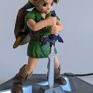 May include: A green-clad Link figurine from The Legend of Zelda series, holding the Master Sword. The figurine is posed in a fighting stance, with the sword raised above his head. The base of the figurine is a gray stone with a Triforce symbol. The figurine is lit from below, casting a blue glow on the base.