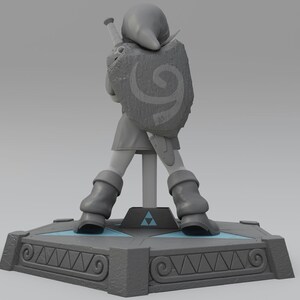 May include: A gray plastic figurine of Link from the Legend of Zelda video game series. Link is wearing a green tunic and holding a sword and shield. The figurine is standing on a gray base with a Triforce symbol.