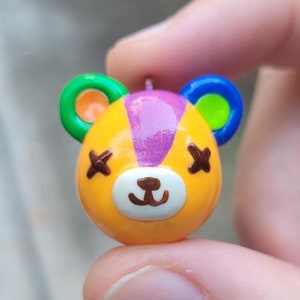 May include: A small, yellow, purple, green, and blue clay figurine of a bear with an X on each eye and a brown nose. The bear is wearing a rainbow colored headband.