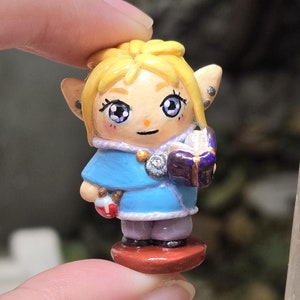 May include: A hand-painted figurine of a young girl with blonde hair, wearing a blue and white dress and holding a purple book. The figurine is standing on a brown base.