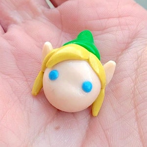 May include: A small, hand-crafted figurine of a cartoon character with blonde hair, green hat, and blue eyes. The figurine is made of polymer clay.