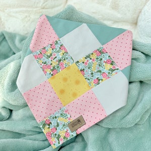 May include: A patchwork dog bandana featuring a floral, polka dot, and sun design in pink, yellow, and blue. The bandana has a brown leather tag with the text "Oh Happy Barkery".